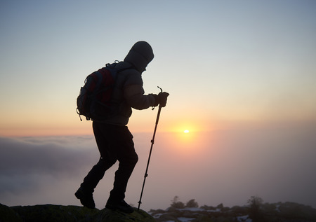 Tourist hiker in warm clothing with backpack and trekking sticks walking on rocky mountain peak on foggy cloudy landscape, misty blue sky and raising bright orange sun at dawn copy space background.の写真素材