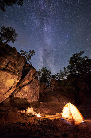 Mountain campsite at summer night amid huge steep rock formation. Small tourist tent brightly lit by burning campfire under clear dark starry sky with Milky way. Tourism, hiking and traveling conceptの写真素材