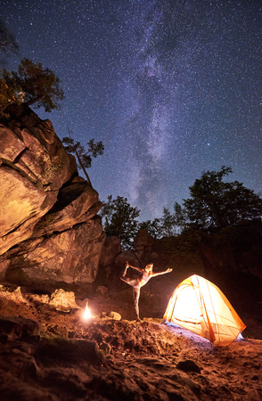 Campsite at night amid huge steep mountain rock formations. Attractive young woman doing yoga exercises at tourist tent on clear dark blue starry sky background. Tourism and sport concept. Narajasanaの写真素材