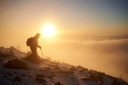 Tourist climber with backpack and trekking poles climbing on rocky snowy mountain steep slope on background of foggy valley filled with white puffy clouds, raising sun and blue sky at sunrise.の写真素材