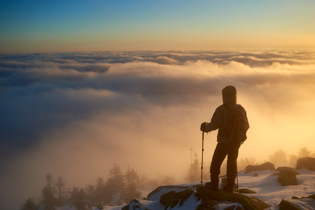 Back view of tourist hiker with backpack and hiking sticks standing on rocky mountain peak, enjoying beautiful view of foggy valley filled with white puffy clouds at sunrise.の写真素材