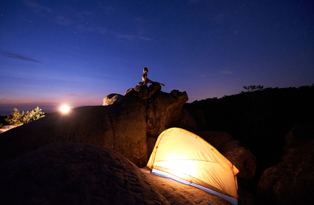 Small silhouette of slim woman doing complicated yoga exercises on top of huge boulder against dramatic dark blue red sky and setting sun. Tourism, active lifestyle, climbing and camping concept.の写真素材