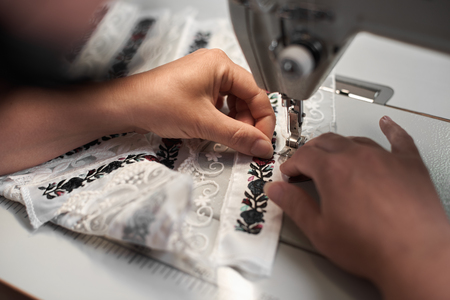 Female seamstress hands connecting white fabric details with floral colorful embroidery with stripes...