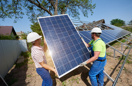 Team of two technicians working on exterior voltaic solar panel system installation in rural countryside on bright sunny summer day. Renewable ecological cheap green energy production concept.の写真素材