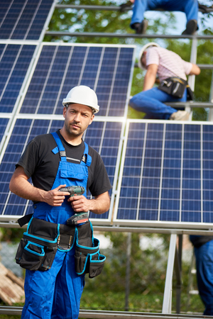 Portrait of male engineer technician with electrical screwdriver standing in front of unfinished high exterior solar panel photo voltaic system blue surface with team of workers on high platform.の写真素材