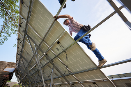 Bottom view from inside of stand-alone exterior solar panel system with engineer installing on high steel platform on bright clear blue summer sky background. Ecological energy production concept.の写真素材