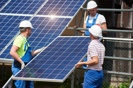 Team of three young technicians in protective helmets mounting heavy solar photo voltaic panel on metal platform. Exterior solar panel system installation, efficiency and durability concept.の写真素材