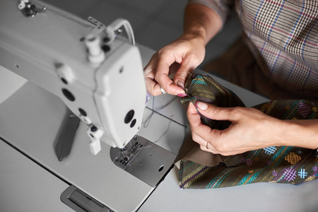 Tailor workplace: view from above of electrical sewing machine and close-up of female hands with needle stitching together garment details before sewing. Handmade exclusive fashionable clothes making.の写真素材