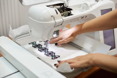 Close-up of woman manicured hands working on sewing machine creating colorful geometrical pattern on white textile detail. Modern technology, fashion and traditional clothing manufacturing concept.の写真素材