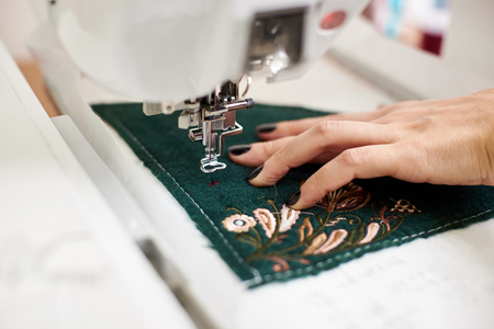 Close-up of female manicured hand working on sewing machine creating colorful floral pattern on dark green textile detail. Modern technology, fashion and traditional clothing manufacturing concept.の写真素材