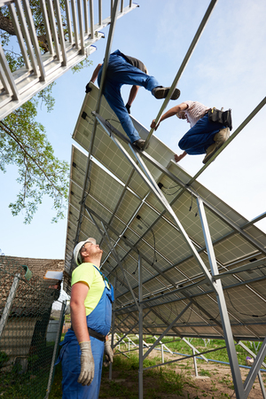 View from inside of stand-alone exterior solar voltaic system. Engineer technician watching team of workers on high steel platform on bright clear blue sky background. Ecological energy production.の写真素材