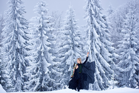 Attractive young slim woman in dark long fantasy outfit and short white sleeveless embroidered coat with raised arm outdoors in deep snow on sunny winter day on background of snowy spruce trees.の写真素材