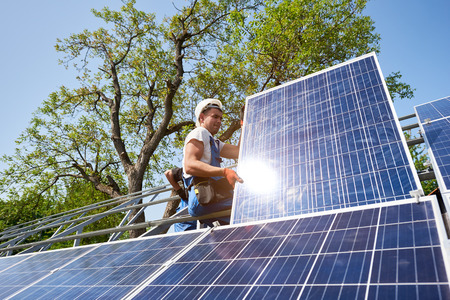 Young technician sitting on metal platform adjusting heavy solar photo voltaic panel on blue sky and green tree background. Stand-alone solar panel system installation and professionalism concept.の写真素材