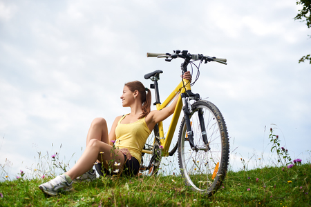 Young happy woman biker sitting on a grassy hill with yellow mountain bicycle, enjoying summer day in the mountains. Outdoor sport activity, lifestyle conceptの写真素材