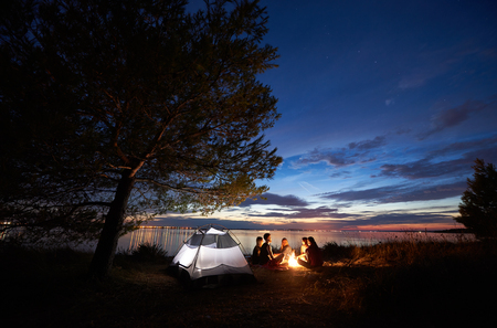 Group of five tourists having a rest on lake shore around campfire near tent under big tree and blue evening sky with first stars at sunset. Tourism, friendship camping and beauty of nature concept.の写真素材
