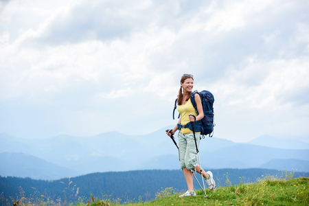 Attractive happy woman tourist hiking mountain trail, walking on grassy hill, wearing backpack, using trekking sticks, enjoying summer cloudy day. Outdoor activity, tourism conceptの写真素材