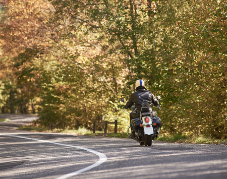 Back view of motorcyclist in black leather jacket and helmet riding cruiser motorbike along twisted asphalt road on bright sunny autumn day on yellow forest trees foliage background.の写真素材