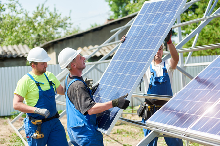 Team of three young technicians in protective helmets mounting heavy solar photo voltaic panel on metal platform in back yard of village house. Cheap ecological green energy provider concept.の写真素材