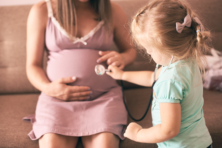 Selective focus of cute daughter playing doctor and checking pregnant mother. Happy woman sitting on sofa at home and playing with lovely girl. Concept of motherhood and happiness.の写真素材