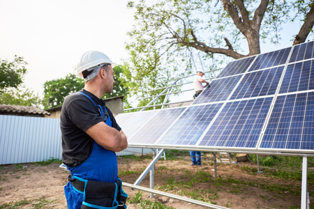 Engineer technician standing in front of unfinished high exterior solar panel photo voltaic system blue shiny surface with team of workers on high platform.の写真素材
