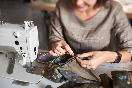 Tailor workplace: view from above of electrical sewing machine and close-up of woman hands with needle stitching together garment details before sewing. Handmade exclusive fashionable clothes making.の写真素材