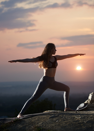 Slim attractive long-haired girl with stretched arms in standing yoga pose outdoors on big boulder on background of cloudy beautiful foggy pink sky and setting over horizon bright sun. Virabhadrasanaの写真素材