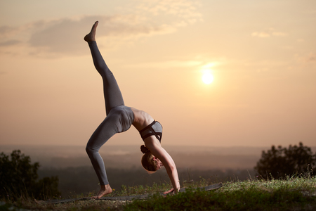 Profile of slim strong muscular girl with raised leg doing complicated yoga exercises on grassy hill on copy space background of bright light summer sky and blurred misty landscape deep below.の写真素材