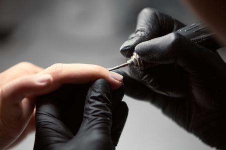 Close-up of professional master beautician hands in black gloves working with electric drill file on client woman fingernails. Finger nail treatment, manicure making in beauty salon.の写真素材
