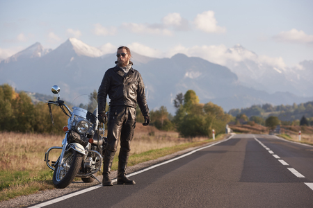 Bearded tall athletic biker in dark sunglasses, black leather clothing standing at modern powerful motorbike on background of green rural landscape, steep mountain peaks and bright sky.の写真素材