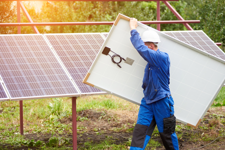 Side view of technician in protective helmet carrying big shiny solar photo voltaic panel on exterior metal platform background on sunny summer warm day. Renewable ecological green energy concept.の写真素材