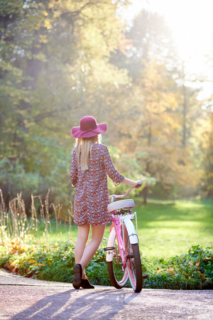 Back view of slim blond fashionable long-haired attractive girl in short dress and pink hat at lady bicycle outdoors on paved summer park alley on beautiful foggy green and golden trees background.の写真素材