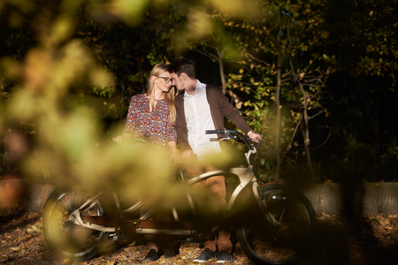 Happy romantic couple, bearded guy and attractive girl close together at tandem double bicycle outdoors in dark autumn park or forest at sunset on blurred golden green bokeh background.の写真素材