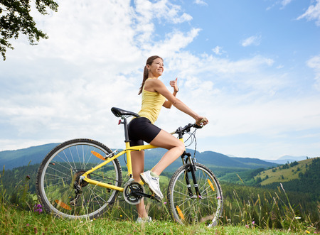 Attractive happy female cyclist riding on yellow mountain bike on a grassy hill, showing thumbs up, enjoying summer day in the mountains. Outdoor sport activity, lifestyle conceptの写真素材
