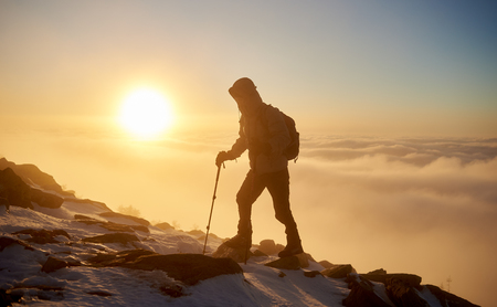 Back view of tourist hiker with backpack and hiking sticks hiking on rocky mountain peak on copy space background of beautiful foggy valley filled with white puffy clouds at dawn at winter.の写真素材