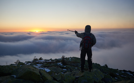 Back view of tourist in warm jacket with backpack on rocky mountain peak pointing with hiking stick at beautiful foggy valley covered with white clouds and bright morning sun rising on horizon.の写真素材