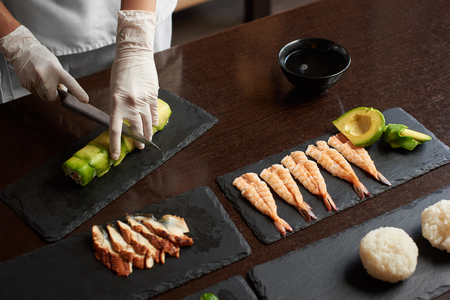 Closeup view of process of preparing rolling sushi. Hands are cutting roll on the black stone plateの写真素材