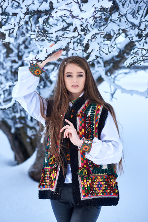 Young beautiful long-haired woman in white embroidered blouse and multicolored vest outdoors on sunny winter day on snowy landscape background. Traditional womens attire of western part of Ukraine.の写真素材