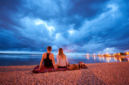Back view of young bearded man and long-haired woman sitting on rug on resort town pebble beach at dusk, enjoying view of cruise boats floating in calm blue water and dramatic cloudy evening skyの写真素材