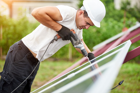 Profile of professional technician working with screwdriver connecting solar photo voltaic panel to metal platform on green summer blurred bokeh background. Alternative sun energy production concept.の写真素材