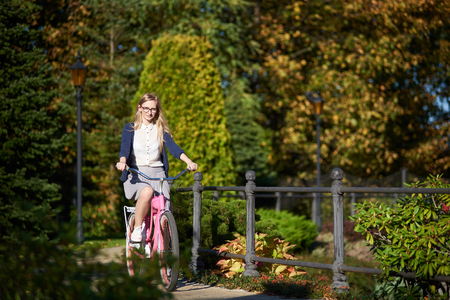 Happy blond long-haired attractive traveler woman in glasses and casual clothing cycling modern pink lady bike along narrow park bridge on bokeh background of beautiful different green trees.の写真素材