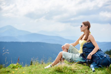 Side view of attractive woman hiker resting on grassy hill with backpack and trekking sticks, wearing sunglasses, enjoying summer cloudy day in the mountains. Outdoor activity, tourism conceptの写真素材