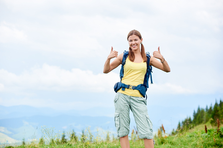 Portrait of attractive smiling woman tourist hiking mountain trail, walking on grassy hill, wearing backpack, using smiling to the camera and showing thumbs up. Outdoor activity, tourism conceptの写真素材