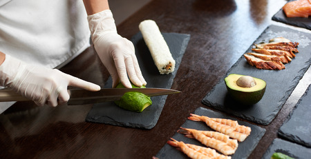Close up view of process of preparing delicious rolling sushi in restaurant. Female hands in disposable gloves slicing avocado on wooden board with the knife.の写真素材