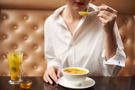 Crop of young female in white eating tasty orange cream soup. Woman in blouse enjoying tasty first dish in luxury restaurant. Client eating exotic meal on lunch. Concept of oriental food.の写真素材
