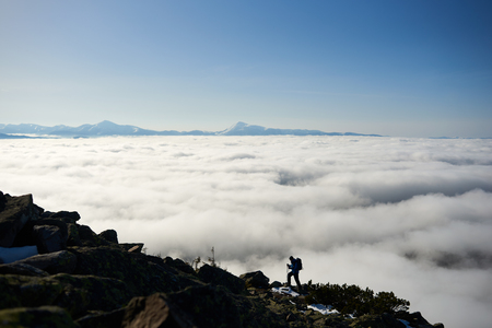 Small silhouette of tourist hiker with backpack and trekking poles climbing steep rocky mountain slope on background of blue sky, foggy valley filled with white clouds and snowy mountain tops.の写真素材