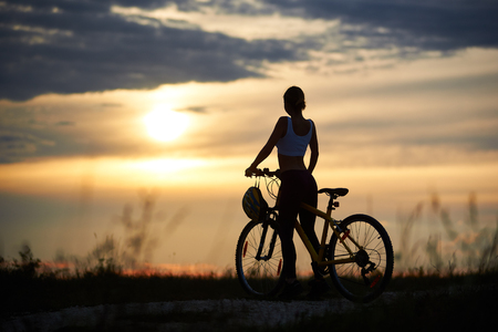 Back view of woman with bicycle standing on road among grass enjoying the sunset on evening sky. The girl has a beautiful sporty body. Helmet hangs on the handle of bicycleの写真素材