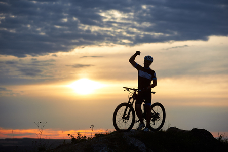 Sporty smiling man stands with bicycle on rock on top of a hill on a blurred background of a beautiful sunset. The man is dressed in sportswear and a helmetの写真素材