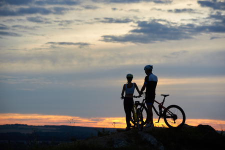 Rear view of two young people with mountain bikes stand on top of a cliff with beautiful scenery of hills and sky at sunset. The concept of a healthy lifestyleの写真素材