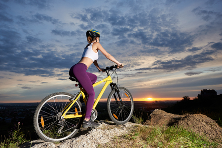 Side view of athletic and sporty girl sitting on her bicycle and holding hands on handle bar. Slender female cyclist posing on hill enjoying great view of landscapes and sunset.の写真素材