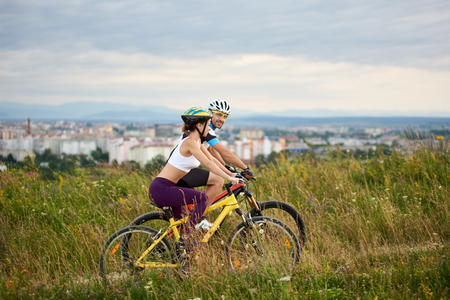 Two positive and energetic cyclists riding bicycle on trail in high grass. Beautiful and cheerful man and woman in sportswear and helmets. Behind them city and built environment.の写真素材